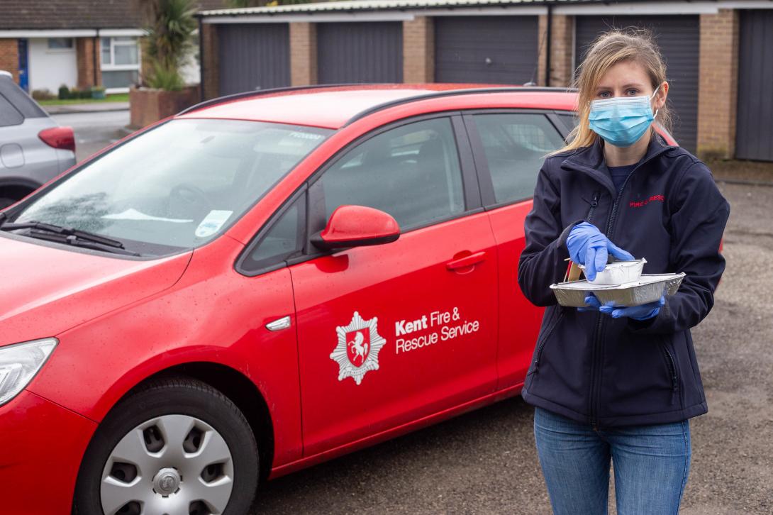 A female KFRS employee with face covering stands next to a red KFRS car while delivering food to customers during the pandemic